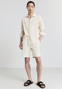 Giovane uomo in piedi che indossa una camicia a maniche lunghe color crema con bottoni, pantaloni corti abbinati e sandali neri aperti di fronte a uno sfondo bianco semplice.