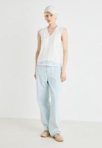 Hvit blondetopp uten ermer med v-hals-design, med delikate bølgede kanter, kombinert med lyseblå løstsittende jeans og beige sko.