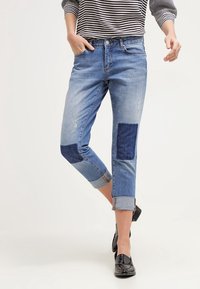 Džínové kalhoty s středním umytím, s ohrnutými manžetami, volným střihem a nášivkami v tmavě modré barvě. Zničené detaily dodávají texturu.