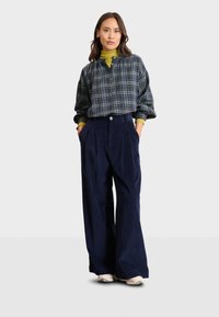 Pantaloni ampi in corduroy blu navy con pieghe frontali, abbinati a un dolcevita verde e una camicia a quadri bottonata con accenti blu e gialli.