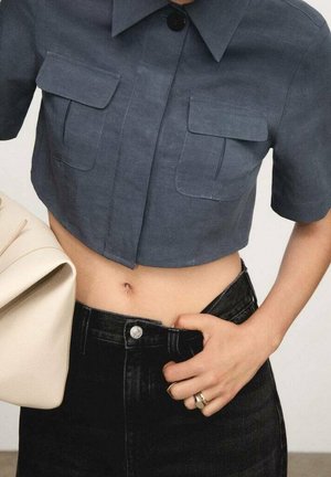 Cropped, dark blue button-up shirt with collar and two chest pockets, paired with high-waisted black jeans, and a light beige bag.
