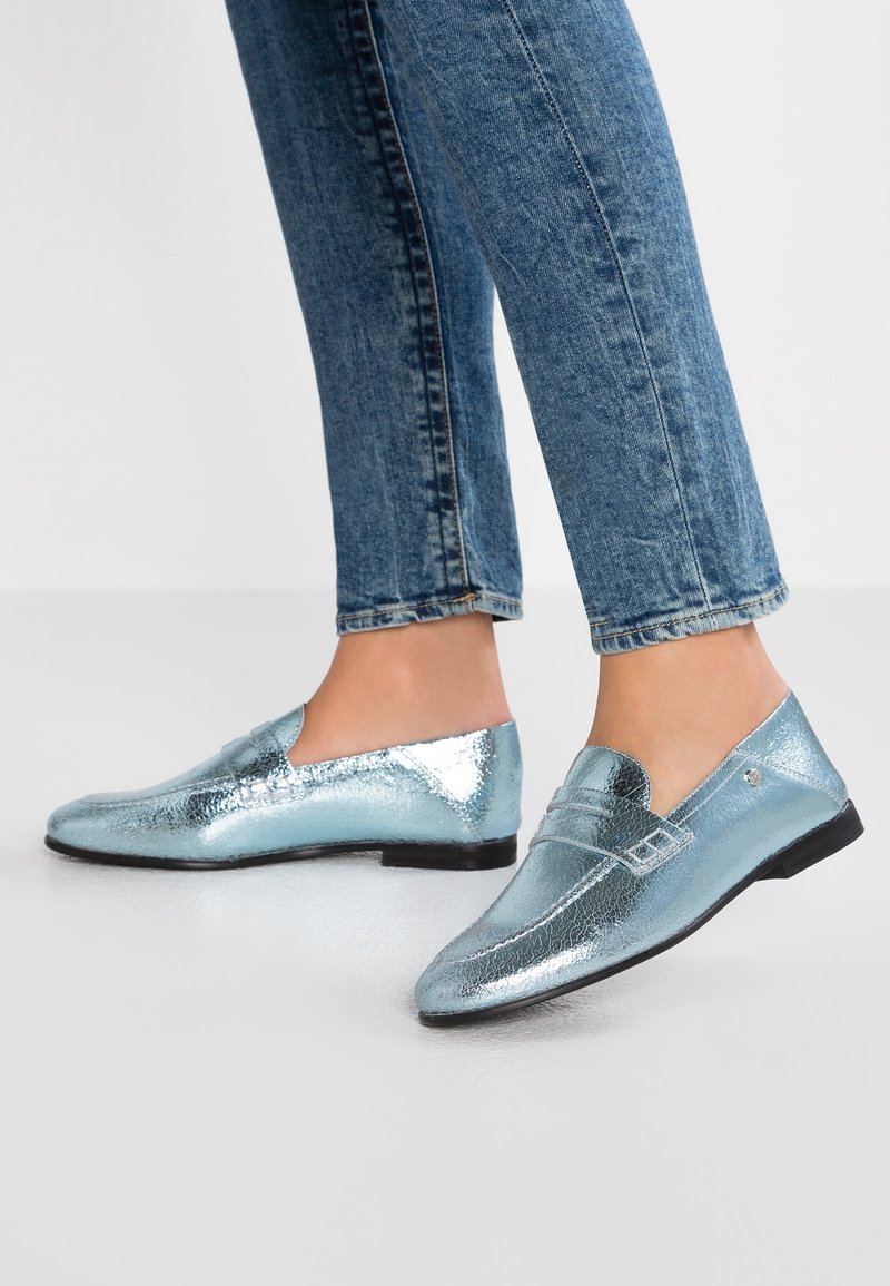 Mocassins bleu clair métallisé avec une texture lisse, un bout arrondi et des détails subtils sur la partie supérieure. Semelle en caoutchouc noire.