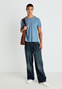 Kék pamut póló kis mellzsebbel, oversize sötét farmerral és fehér sneaker-rel párosítva, egy barna válltáskával a kezében.