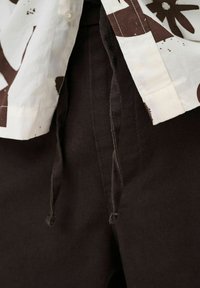 Pantalon noir à cordon avec extrémités nouées, partiellement couvert par une chemise blanche avec des motifs abstraits foncés et un poignet boutonné.