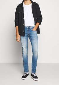 Camisa negra abotonada sobre camiseta blanca, combinada con vaqueros azul claro con rotos en las rodillas y zapatillas negras con suelas blancas.