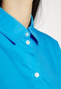 Blue collared shirt with shiny texture, featuring white buttons and visible stitching along the placket. The collar is pointed and structured.