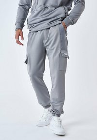 Grå joggebukser med en glat finish, der har en sidelomme til cargo, elastiske manchetter og hvid branding. Sammen med hvide sneakers.