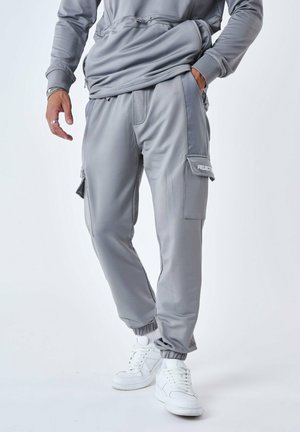 Grå joggebukser med en glat finish, der har en sidelomme til cargo, elastiske manchetter og hvid branding. Sammen med hvide sneakers.