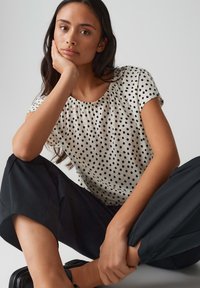 Korte mouwen blouse in crème met zwarte stippen, ronde halslijn, geplooide details, gecombineerd met losse zwarte broeken.