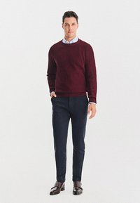 Burgundowy sweter o prążkowanej fakturze, niebieska koszula z kołnierzykiem pod spodem, w połączeniu z ciemnogranatowymi spodniami i brązowymi skórzanymi butami. Neutralne tło.