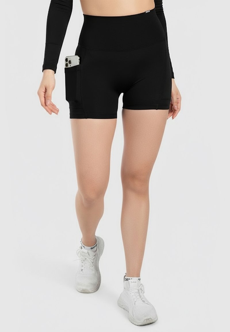Femme portant un short cycliste noir taille haute avec une poche latérale tenant un smartphone, des baskets blanches et un haut noir à manches longues.
