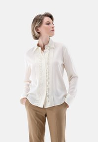 White blouse with button-down collar, vertical pleats, and ruffled front. Soft fabric, long sleeves, paired with beige trousers.