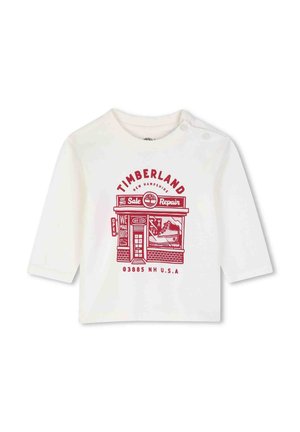 T-shirt blanc à manches longues pour tout-petit avec un graphisme rouge du magasin Timberland et un texte, présentant un design de botte et d'atelier de réparation à l'avant.