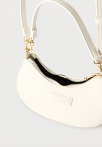 Μικρή λευκή δερμάτινη τσάντα Lacoste με χρυσλό φερμουάρ και ρυθμιζόμενο λουρί, με ελάχιστες ραφές και μπροστινή ετικέτα με το λογότυπο.