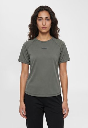Vrouw met lang golvend haar, gekleed in een grijs sportshirt met korte mouwen en zwarte broek, staand tegen een effen achtergrond.