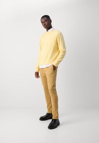 Pull-over jaune clair à manches longues, superposé à une chemise à col blanc, associé à un pantalon beige et des chaussures noires, avec une texture lisse.