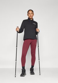 Pullover nero con zip, leggings bordeaux, scarpe da trekking nere. Il modello tiene in mano dei bastoncini da trekking. Tessuto liscio con dettagli del logo e design aderente.