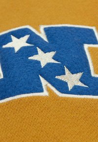 Gelber Stoff mit einem blauen, filzigen "N", verziert mit weißen Sternen. Die Nähte sind sichtbar und betonen die Textur und Farben.