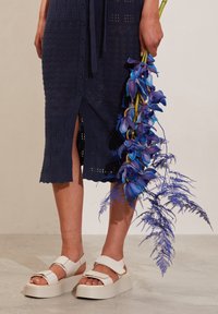 Persona in una gonna midi a maglia blu navy e sandali con platform bianchi tiene un lungo stelo di orchidee blu vibranti con foglie di felce.
