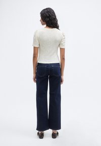 Ανοιχτόχρωμο, πλεκτό τοπ με υφή και φουσκωτά μανίκια, σε συνδυασμό με σκούρο μπλε τζιν παντελόνι φαρδιάς γραμμής. Τα παπούτσια έχουν καφέ δερμάτινες λεπτομέρειες.