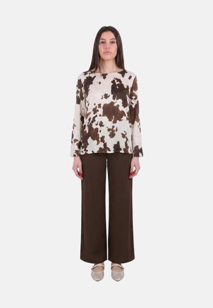 Donna in piedi, indossa una maglia beige a maniche lunghe con stampa astratta marrone, pantaloni ampi marrone scuro e ballerine beige con cinturini alla caviglia.