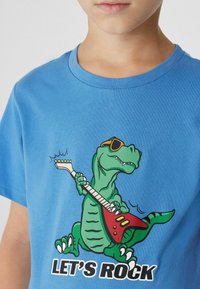Blaues Baumwoll-T-Shirt mit einem Grafikdesign eines grünen Dinosauriers mit Sonnenbrille, der eine rote E-Gitarre spielt, der Text lautet "LASST UNS ROCKEN."