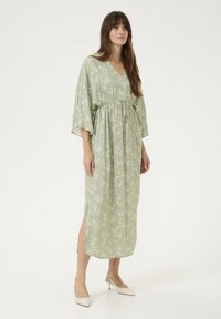 Cvečni, svetlo zeleni maksi obleki z V-izrezom, zategnjeno pasom in širokimi rokavi. Ima stranske razpoke in tekočo teksturo blaga.