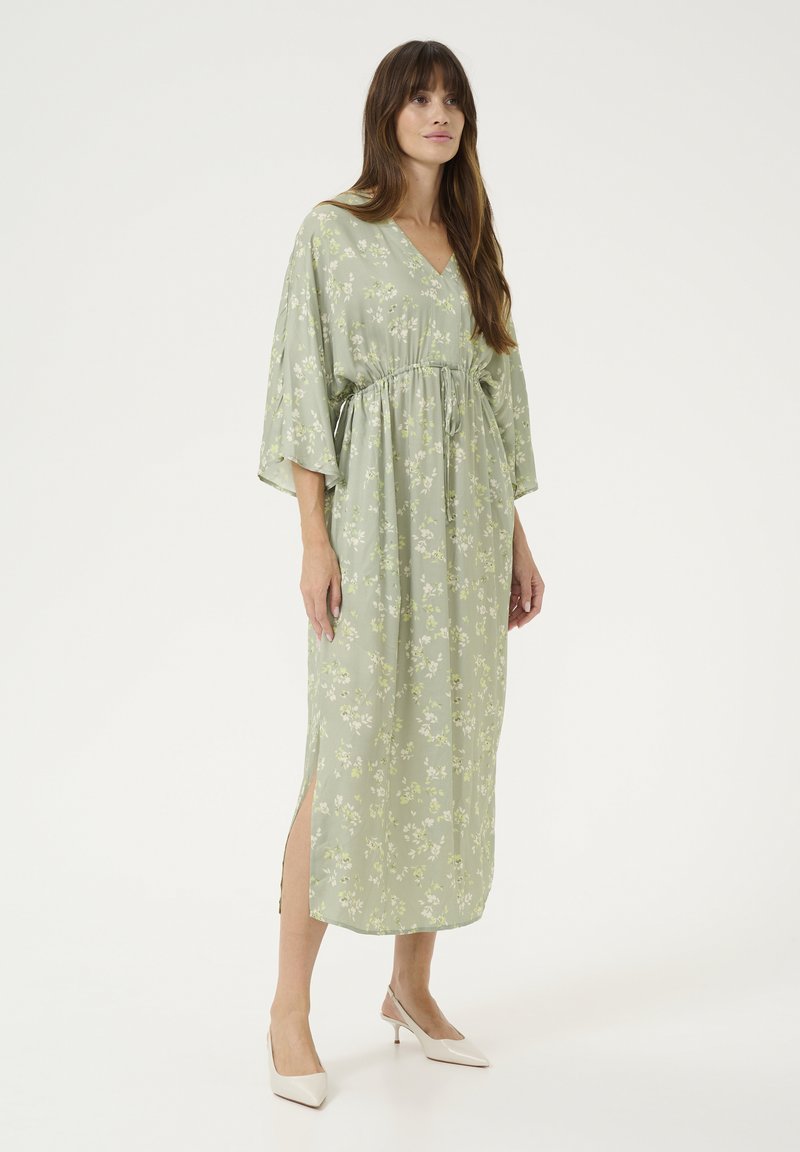 Cvečni, svetlo zeleni maksi obleki z V-izrezom, zategnjeno pasom in širokimi rokavi. Ima stranske razpoke in tekočo teksturo blaga.