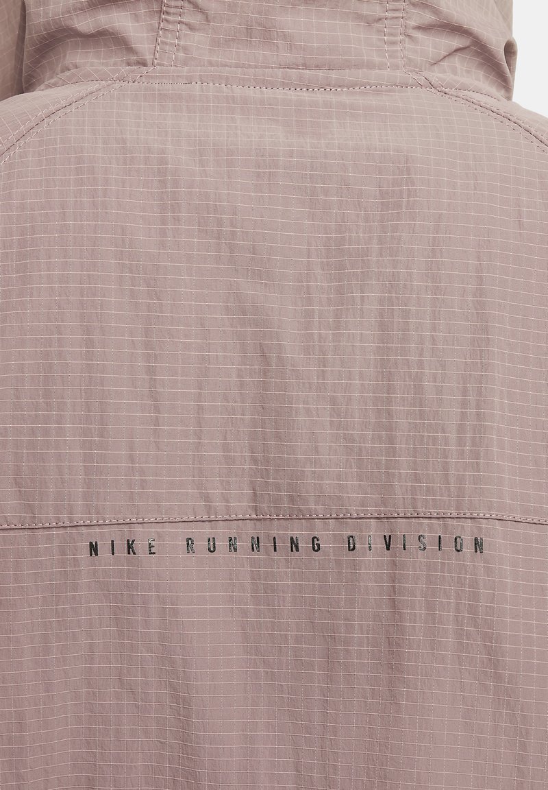 Nike Running Division-jack in lichtroze, met een ruitpatroon, een gladde textuur en zwarte letters op de achterkant.