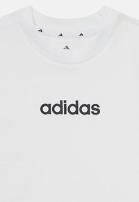 Valkoinen puuvillapaita, jossa on musta painettu "adidas"-logo keskellä, ribbireunuksella varustettu pyöreä kaula-aukko ja sileä pinta.