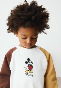 Valge dressipluus pruunide ja vaigistatud kollaste raglanvarrukatega, millel on tikitud Mickey Mouse'i graafika ja tekst all.