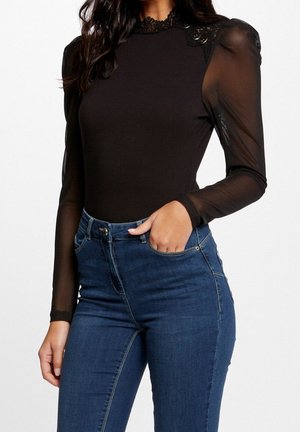 Femme portant un haut noir à manches longues avec manches transparentes et détails en dentelle, associé à un jean en denim bleu taille haute.