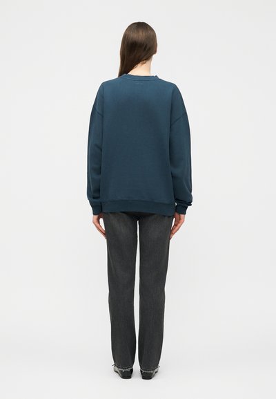 Kvinne står med ryggen mot, iført en teal genser, mørkegrå jeans med rette ben og svarte sko på hvit bakgrunn.