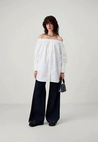 Camicia bianca a spalle scoperte con maniche lunghe, chiusura frontale con bottoni; abbinata a pantaloni larghi blu navy e scarpe con piattaforma nere; piccola borsa nera.