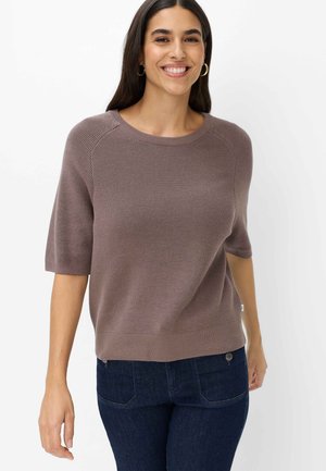Donna sorridente che indossa un maglione marrone a maniche corte e jeans blu scuro su uno sfondo bianco semplice.