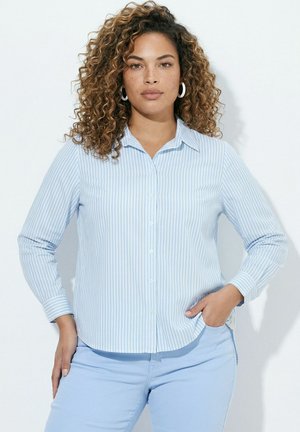 Kvinde med krøllet hår iført hvid og lyseblå stribet skjorte og lyseblå bukser, står med hænderne på hoften.