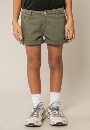 Personne portant un short olive retroussé, des chaussettes blanches montantes et des baskets beige avec des lacets gris, debout sur un fond neutre.