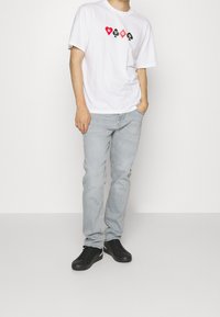 T-shirt bianca in cotone con design grafico nero e rosso che legge "HUGO", abbinata a jeans blu chiaro e sneakers nere.