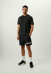 Melna t-kreklu un tumši zilos šortus ar "NIKE" logotipu, kas apvienoti ar melniem sporta apaviem un baltām pusgarajām zeķēm. Modelis stāv neitrālā fonā.