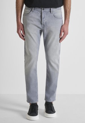 Uomo che indossa jeans slim fit grigio chiaro e sneakers basse nere con suole bianche, in piedi contro uno sfondo chiaro e uniforme.