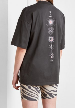 Osoba nosząca ciemny, oversizowy T-shirt z pionowym niebiańskim wzorem na plecach oraz beżowe spodenki w wzór zebry.