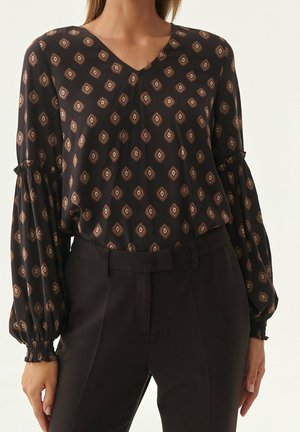 Frau trägt eine schwarze V-Ausschnitt-Bluse mit beigen Diamantmustern und gerafften Manschetten, kombiniert mit hochtaillierter schwarzer, maßgeschneiderter Hose.