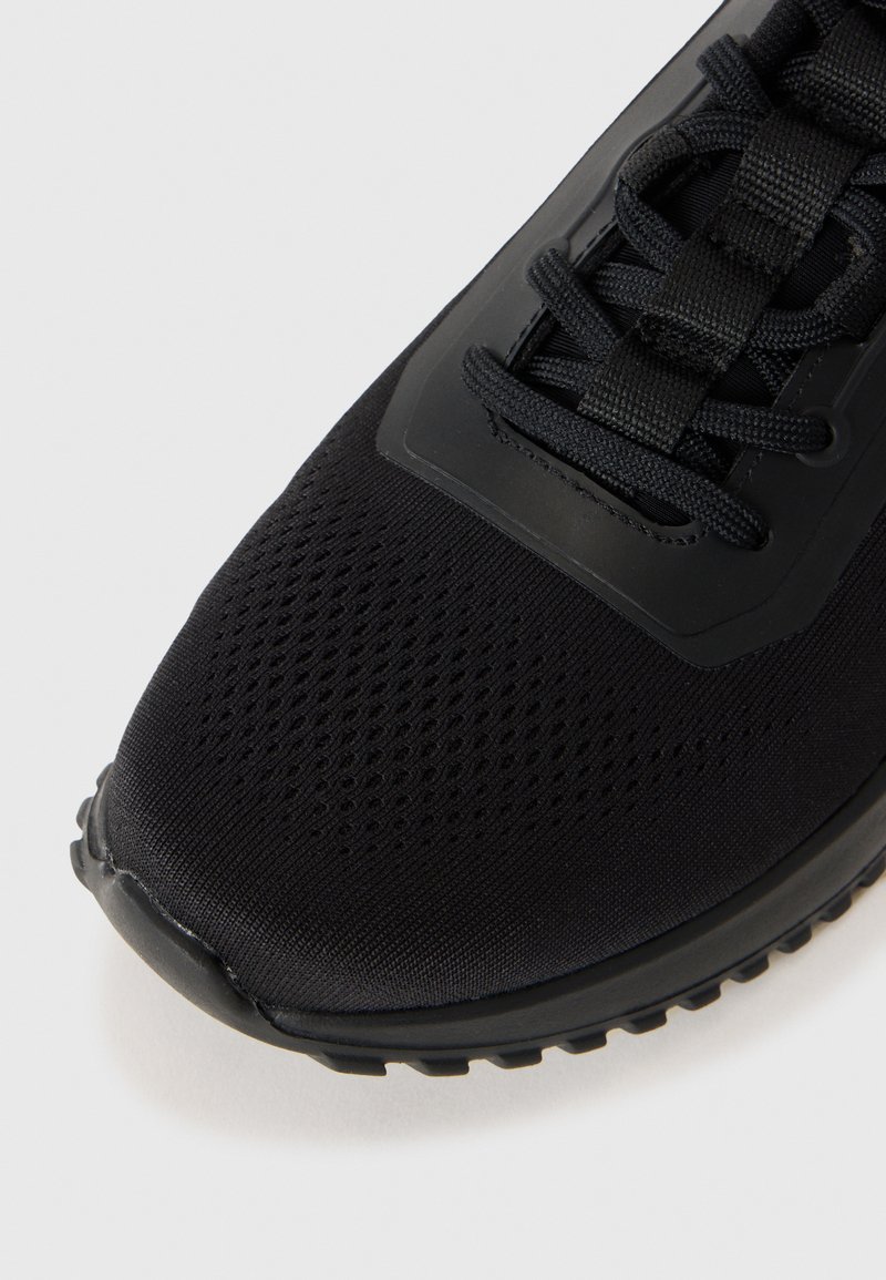 Zapatilla deportiva negra con parte superior de malla, puntera reforzada, área de cordones texturizada y suela de goma flexible. Presenta un diseño minimalista y color monocromático.