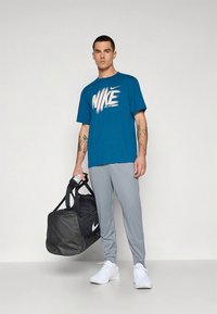Blågrön Nike T-shirt med vitt logo, grå träningsbyxor och vita sneakers. Svart sportväska med vitt logo, hålls i vänster hand.