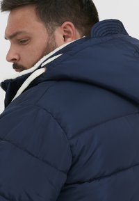 Mørkeblå puffy-jakke med fleeceforet krave og snørehuv. Har kviltet tekstur og kontrasterende hvide detaljer.