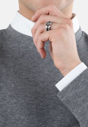 Suéter gris con ribetes y una camisa blanca con cuello debajo. Anillo plateado con un diseño geométrico en el dedo del modelo. Toma de cerca.