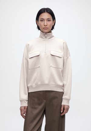 Kobieta z krótkimi ciemnymi włosami ubrana w beżową bluzę na zamek z kieszeniami z klapami oraz luźne brązowe spodnie, stoi na tle jednolitego białego tła.