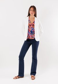 Blazer bianco su una blusa colorata e fantasia con vortici di rosso, arancione e blu; abbinato a pantaloni blu navy e sandali marroni.