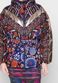 Femme portant une veste colorée à motifs floraux, géométriques et dorés métalliques, zippée et ceinturée à la taille.