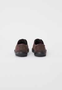 Hnědé suede boty s měkkou texturou, zaobleným tvarem a černými gumovými podešvemi. Pohled z zadní strany ukazuje minimalistický design a žádné viditelné kovové prvky.